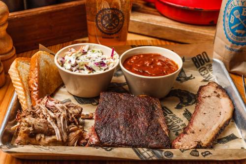 A metal tray with pulled pork, brisket, coleslaw, baked beans, and toasted bread, set on a wooden table.