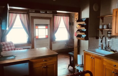 Cozy kitchen with wooden cabinets, a sink, and a small dining area, featuring large windows with checkered curtains.