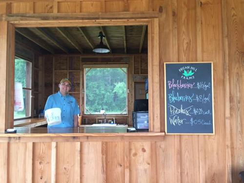 A man stands behind a wooden counter at Pecan Hill Farms, with a chalkboard listing prices for berries and water.