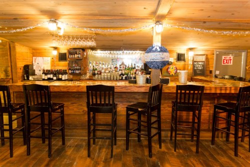 A rustic bar with wooden stools, shelves of bottles, and festive decorations in a cozy, warmly lit setting.