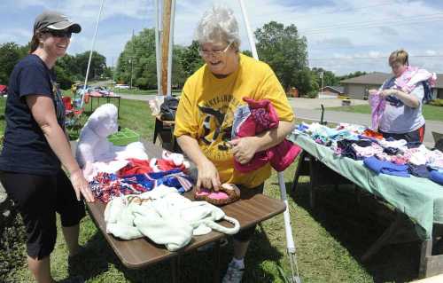 Two women browse items at a yard sale, with clothes and toys displayed on tables in a sunny outdoor setting.