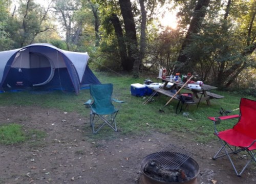 A campsite with a tent, chairs, a picnic table with food, and a fire pit surrounded by trees at sunset.