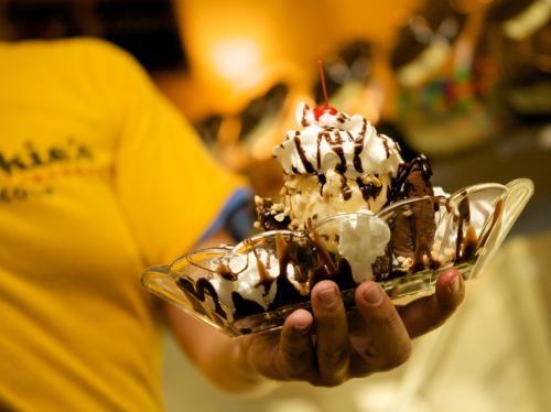 A person holds a large ice cream sundae topped with whipped cream, chocolate sauce, and a cherry in a glass dish.
