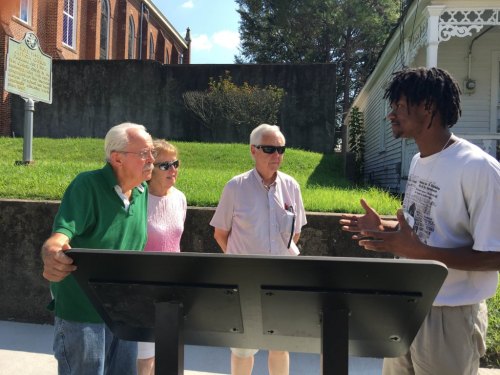 A group of four people engaged in conversation outdoors, with a sign and greenery in the background.