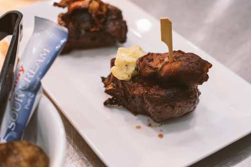 A plated dish featuring grilled meat topped with a small piece of fried food and a toothpick, served on a white plate.