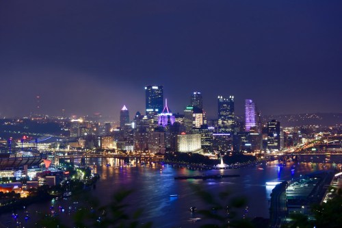 A nighttime view of Pittsburgh's skyline, illuminated buildings reflecting on the river, with bridges in the foreground.