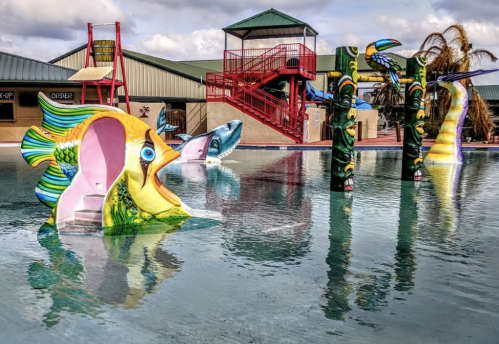Colorful fish sculptures in a pool with a red slide and buildings in the background under a cloudy sky.