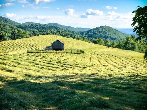 A rustic barn sits on a hillside, surrounded by freshly cut hay and rolling green mountains under a blue sky.