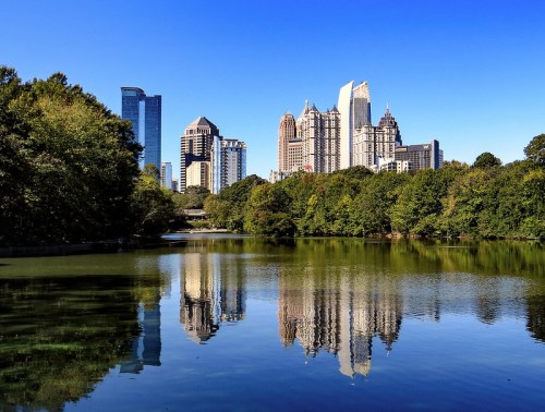 City skyline reflecting on a calm lake, surrounded by lush greenery under a clear blue sky.