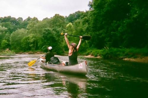 Two people in a canoe on a river, one raising a paddle triumphantly, surrounded by lush green trees.