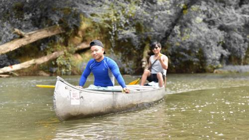 Two people in a canoe on a calm river, surrounded by greenery, enjoying a sunny day outdoors.