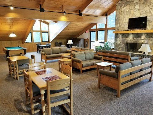 Cozy lounge area with wooden furniture, a stone fireplace, pool table, and large windows letting in natural light.