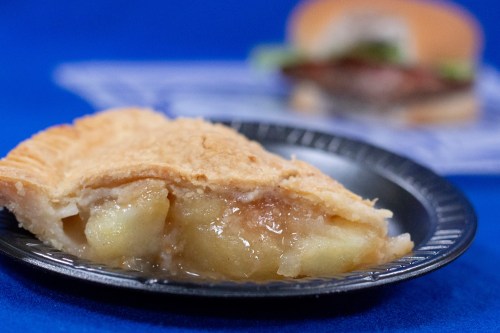 A slice of apple pie on a black plate, with a blurred sandwich in the background.
