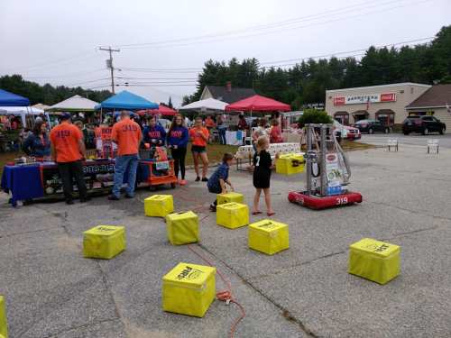 A community event with tents, people, and a robot on display, while children interact with it on a paved area.