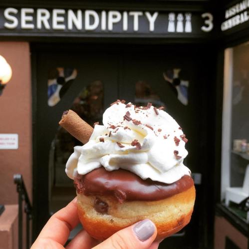 A hand holding a chocolate-covered donut topped with whipped cream and a chocolate stick in front of a shop entrance.