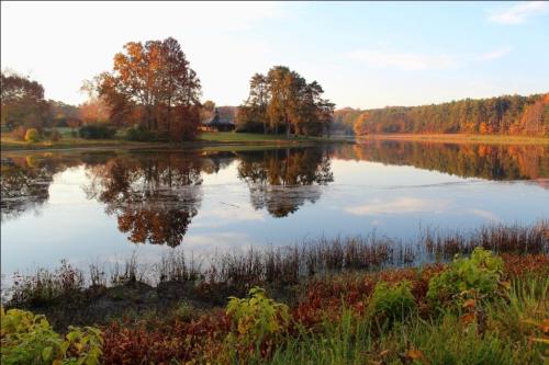 A serene lake reflects autumn trees and a clear sky, surrounded by colorful foliage.
