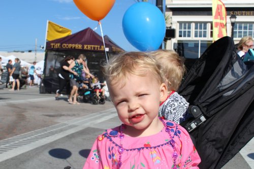 A smiling child in a pink dress holds balloons at a lively outdoor event with people and stalls in the background.