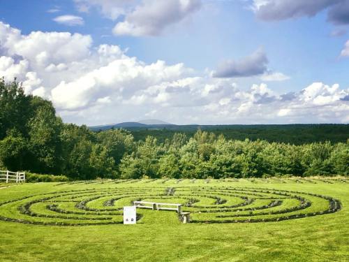 A stone labyrinth set in a green field, surrounded by trees and mountains under a cloudy sky.