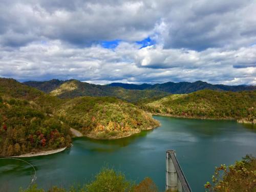 A scenic view of a lake surrounded by colorful autumn foliage and rolling hills under a cloudy sky.