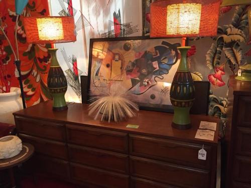 A vintage dresser with two colorful lamps, a decorative object, and an abstract painting in a vibrant setting.