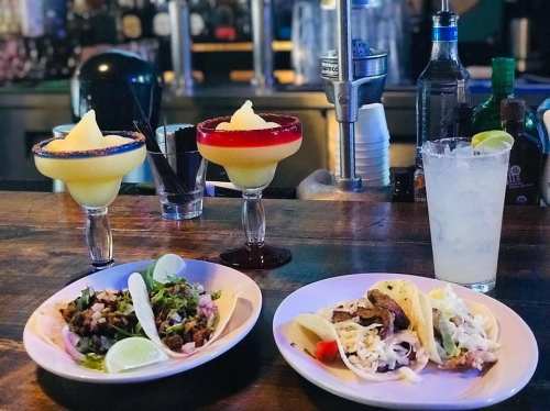 Two colorful cocktails and a plate of tacos with a side of salad and lime wedges on a bar counter.