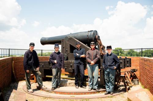 Five men in historical military uniforms stand beside a large cannon on a fortification under a blue sky.