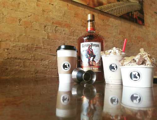 A bottle of Captain Morgan rum next to coffee cups and dessert drinks on a wooden table with a brick wall background.
