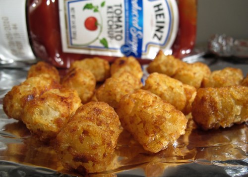 A close-up of crispy tater tots on foil, with a bottle of Heinz ketchup in the background.