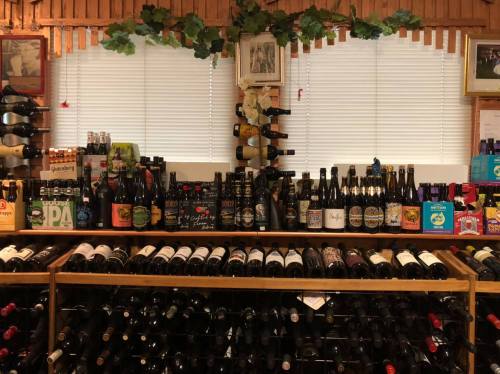 A wooden shelf filled with various bottles of wine and beer, with greenery and framed pictures in the background.
