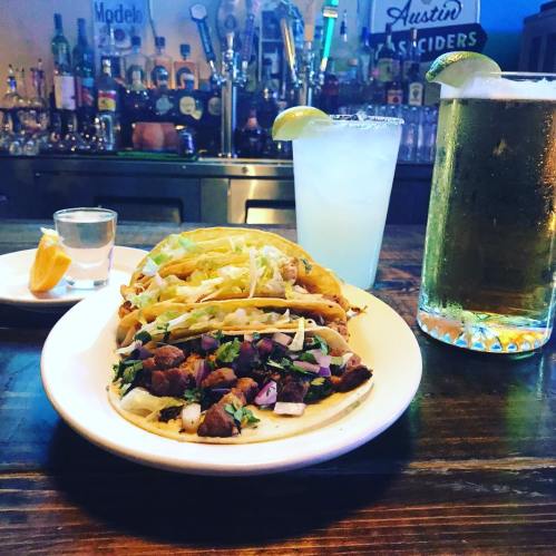 A plate of tacos with toppings, a margarita, a shot of tequila, and two tall glasses of beer on a bar table.