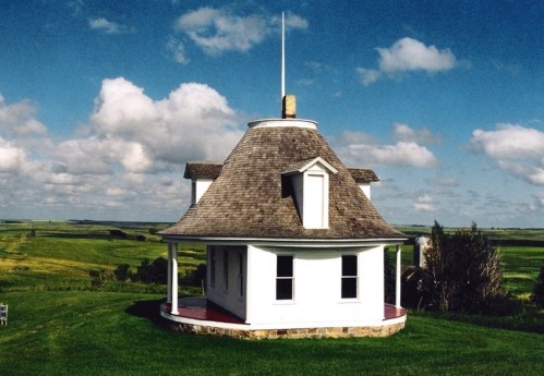 A unique white octagonal house with a conical roof, set against a backdrop of green fields and blue skies.