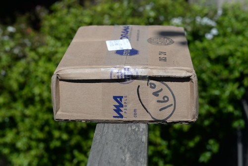 A cardboard box with shipping labels resting on a wooden surface, surrounded by greenery.