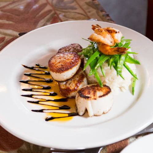 A plate of seared scallops with shrimp, served on rice and topped with green beans and drizzled sauces.