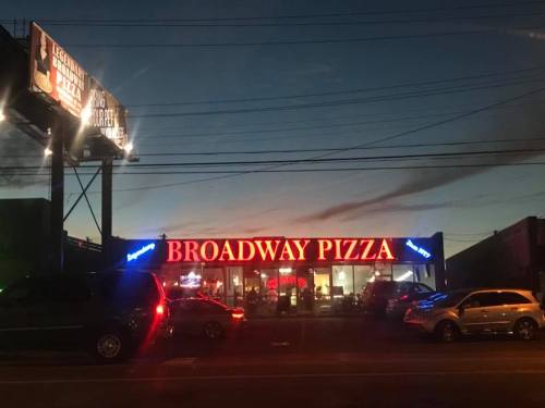 A pizza restaurant named "Broadway Pizza" illuminated at dusk, with cars parked outside and a billboard in the background.