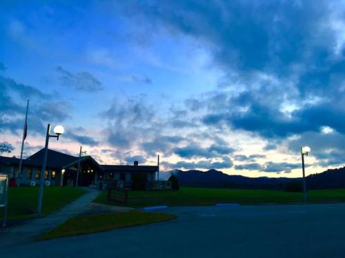 A building with lights stands against a dramatic sky filled with clouds at dusk, surrounded by green fields and mountains.