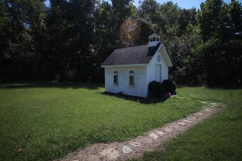A small white building with a steeple sits in a grassy area, surrounded by trees and a stone pathway.