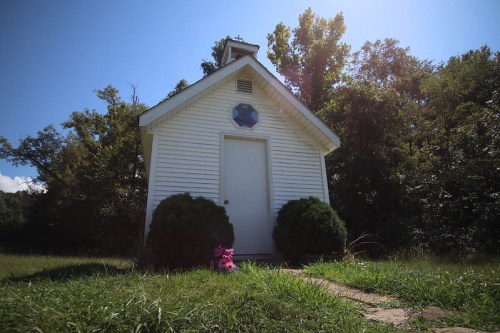 A small white chapel with a star symbol on the front, surrounded by greenery and a clear blue sky.
