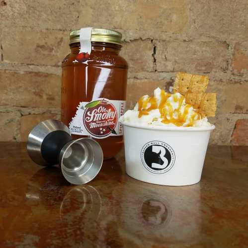 A jar of Ole Smoky Moonshine next to a cup of dessert topped with whipped cream and caramel, with a jigger in the foreground.