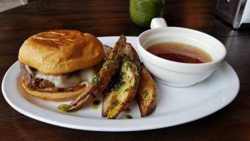 A plate featuring a beef sandwich with melted cheese, roasted potato wedges, and a small bowl of dipping sauce.