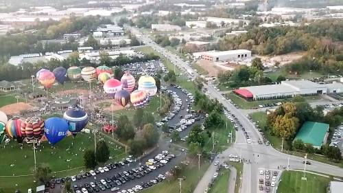Aerial view of a hot air balloon festival with colorful balloons, a crowded parking area, and surrounding greenery.