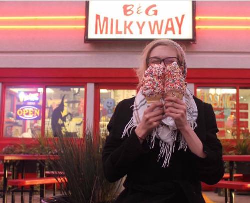 A person holds two large ice cream cones with sprinkles in front of a brightly lit ice cream shop.