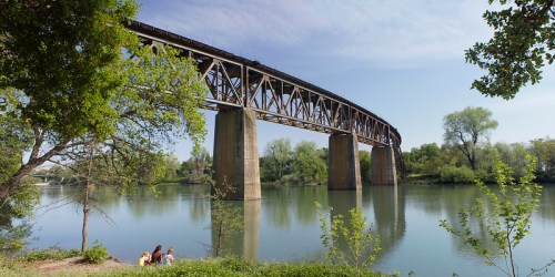 A large metal bridge spans a calm river, surrounded by lush greenery and a few people sitting by the water's edge.