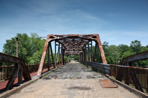 An old, rusty metal bridge spans a quiet landscape, surrounded by trees under a clear blue sky.