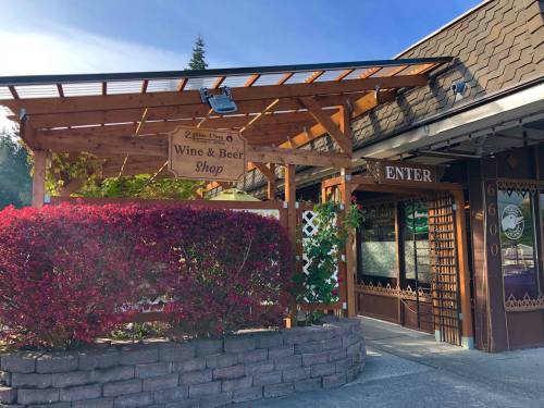 A cozy wine and beer shop entrance with a wooden sign and vibrant red bushes in front.