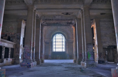 Abandoned interior of a grand building with tall columns, a large window, and graffiti on the walls.