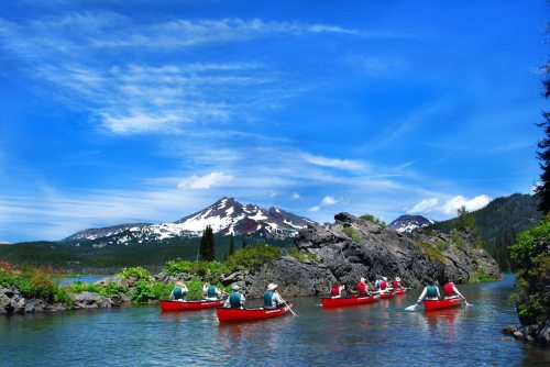 A group of people in red canoes paddling on a serene lake, surrounded by mountains and lush greenery under a blue sky.