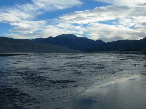 A serene landscape featuring a river flowing through sandy dunes, with mountains and a cloudy sky in the background.