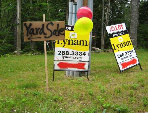 A yard sale sign with balloons next to two real estate signs for Lynam, indicating a sale lot.