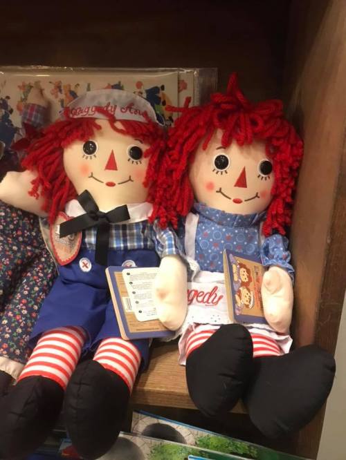 Two Raggedy Ann dolls with red yarn hair, dressed in colorful outfits, sitting side by side on a shelf.