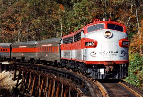 A vintage red and silver train curves around a track surrounded by autumn foliage.